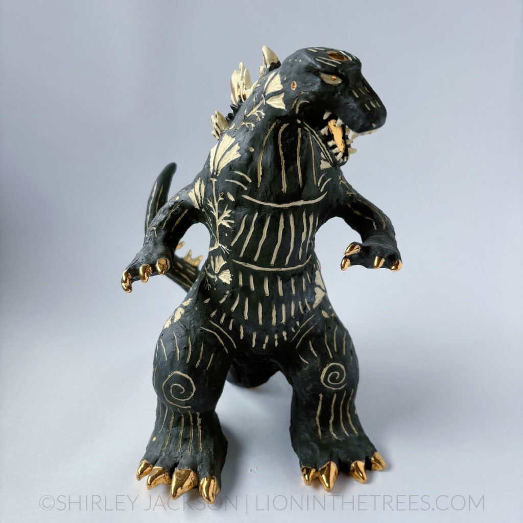 A black sgraffito Godzilla sculpture with California Poppy motifs carved all around it's body. There are gold overglaze details on his spines, claws, and toes.