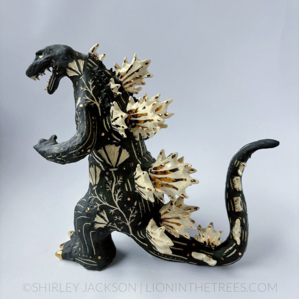 A black sgraffito Godzilla sculpture with California Poppy motifs carved all around it's body. There are gold overglaze details on his spines, claws, and toes.