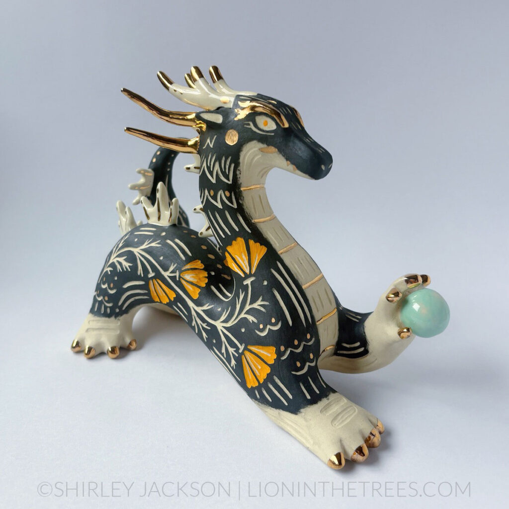 Large, sgraffito eastern dragon sculpture. Done with black and orange underglazes, and gold overglaze details. They are holding a pastel turquoise orb in their left hand.