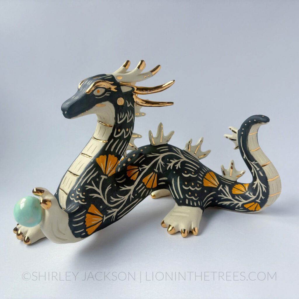 Large, sgraffito eastern dragon sculpture. Done with black and orange underglazes, and gold overglaze details. They are holding a pastel turquoise orb in their left hand.