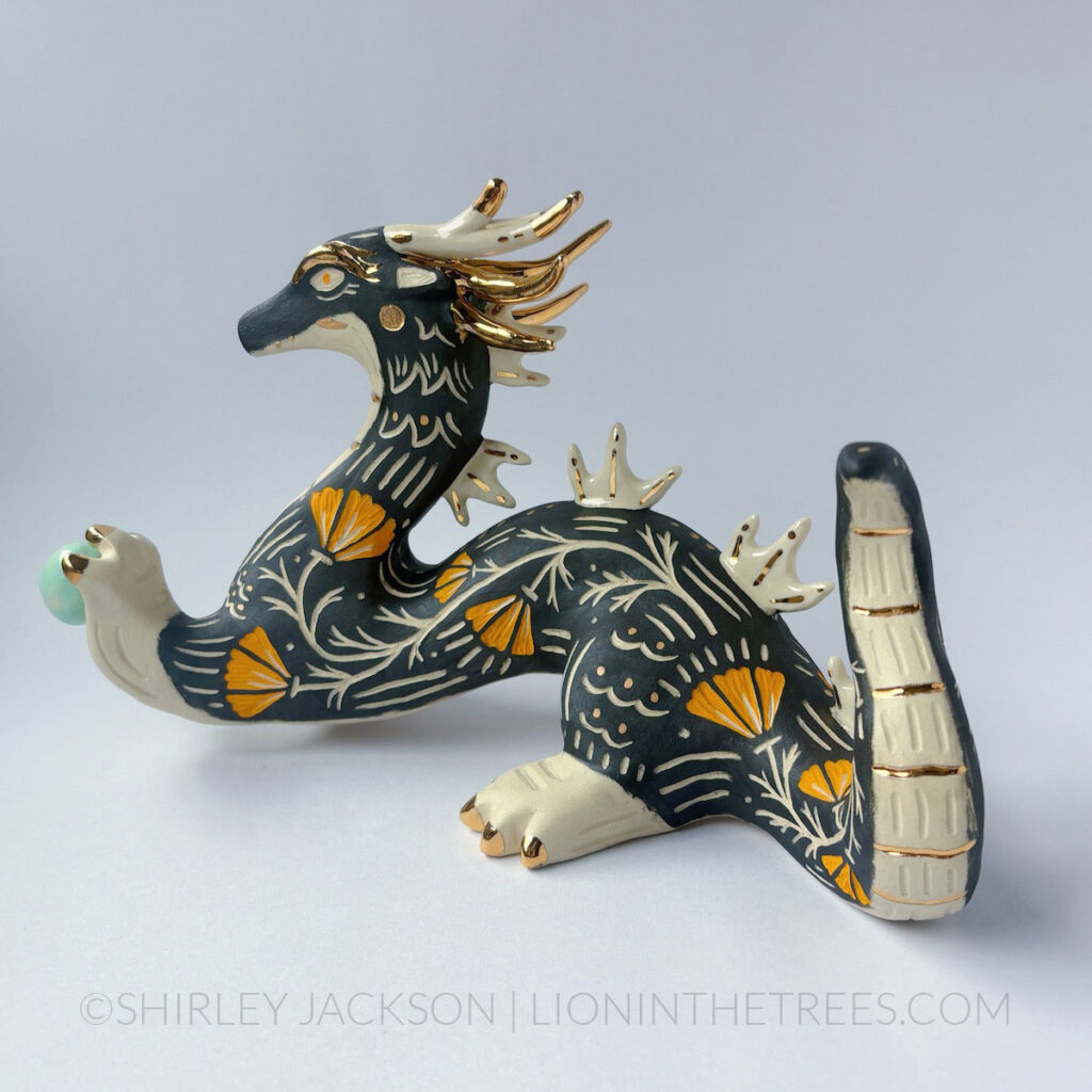 Large, sgraffito eastern dragon sculpture. Done with black and orange underglazes, and gold overglaze details. They are holding a pastel turquoise orb in their left hand.