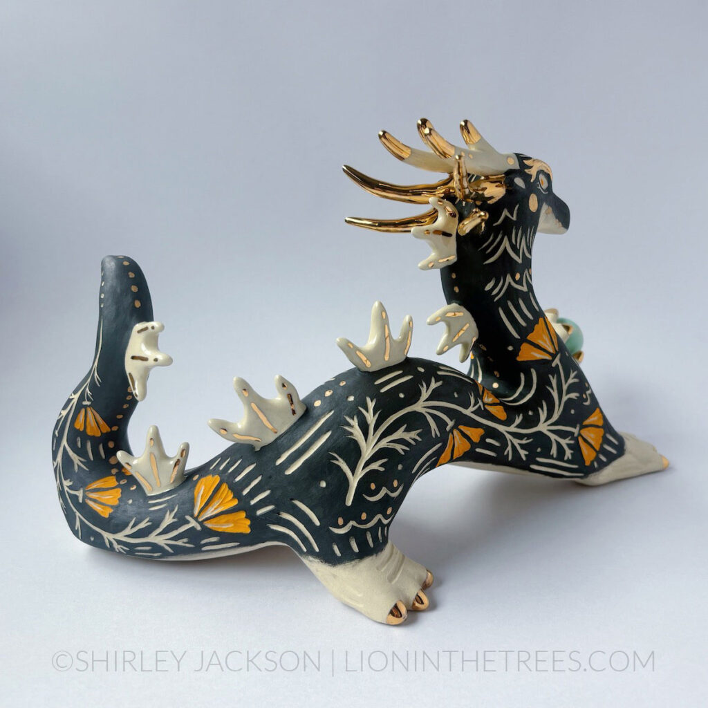 Large, sgraffito eastern dragon sculpture. Done with black and orange underglazes, and gold overglaze details. They are holding a pastel turquoise orb in their left hand.