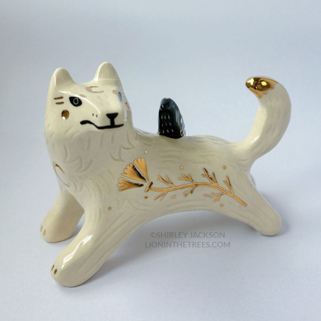 White wolf totem in an outstretched, running position done with minimal black underglaze, finished with clear glaze and gold overglaze details such as a crescent moon on it's chest, forehead, and gold California Poppy motifs on it's side. There is also a small, black raven sitting on their back.