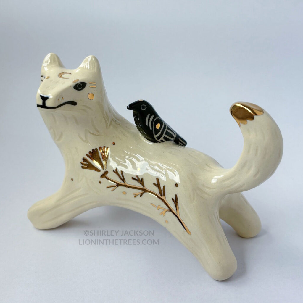 White wolf totem in an outstretched, running position done with minimal black underglaze, finished with clear glaze and gold overglaze details such as a crescent moon on it's chest, forehead, and gold California Poppy motifs on it's side. There is also a small, black raven sitting on their back.