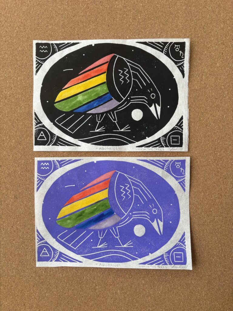 Photo showing two block prints of my rainbow crow Aquarius design. The top print was done with black ink and the print on the bottom was done with the custom lilac ink. The design features watercoloured rainbow wings