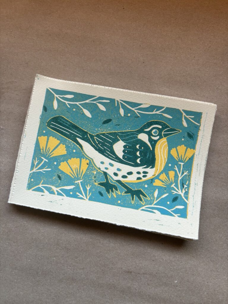 Linoblock print featuring a Yellow-Throated Warbler. It's a small bird with a bright, yellow chest and grey/black/white feathers. The way I've depicted it was using a golden yellow, turquoise blue, and a darker blue/green ink colour. The bird in the design is surrounded by California Poppies on the bottom of the design, and floral leaves/vines creating a border at the top of the print.