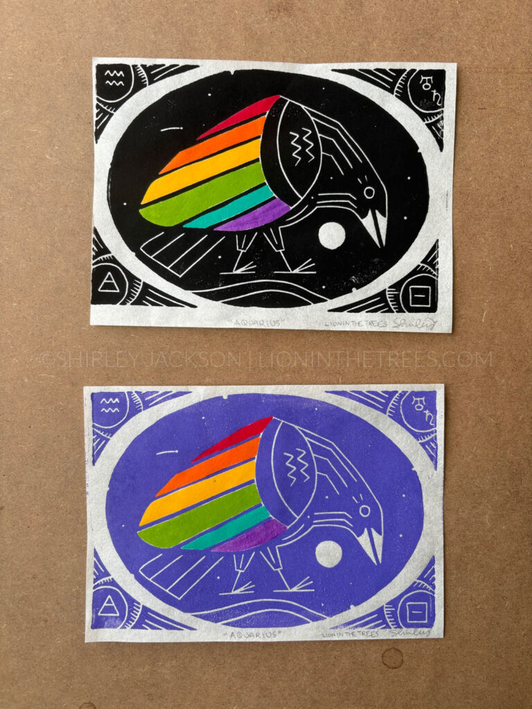 Photo showing two block prints of my rainbow crow Aquarius design. The top print was done with black ink and the print on the bottom was done with the custom lilac ink. The design features acrylic painted rainbow wings so it looks less splotchy.