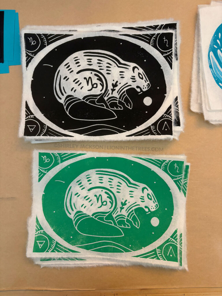 Photo showing two block prints of my beaver capricorn design. The top print was done with black ink and the print on the bottom was done with the green ink.