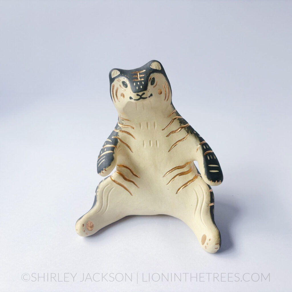 A ceramic tiger sculpture done with a cream white clay and black underglaze with sgraffito markings all over it's body. This shows the front of the piece. The tiger is in a seated-up position almost similar to a human sitting down with their legs splayed out in front of them.