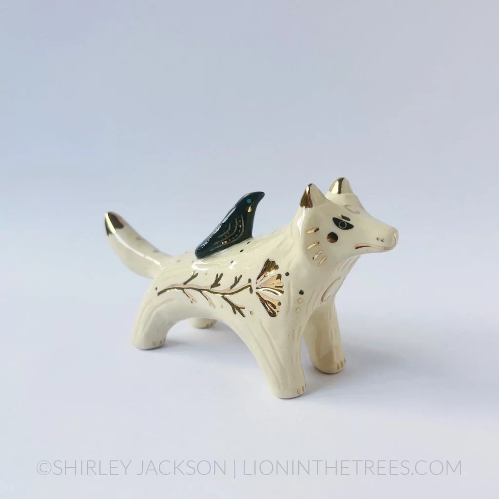 A white and gold ceramic wolf sculpture with gold luster details and has a black raven perched atop it's back.