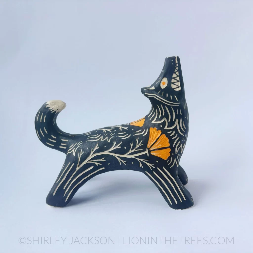 A sgraffito wolf sculpture done in a pose where their neck is arched up, legs stretched up and tail curled as though the wolf was howling up at the moon with a big, toothy grin. They are done with a black and orange underglaze and feature my signature California Poppy motif. Right side view.