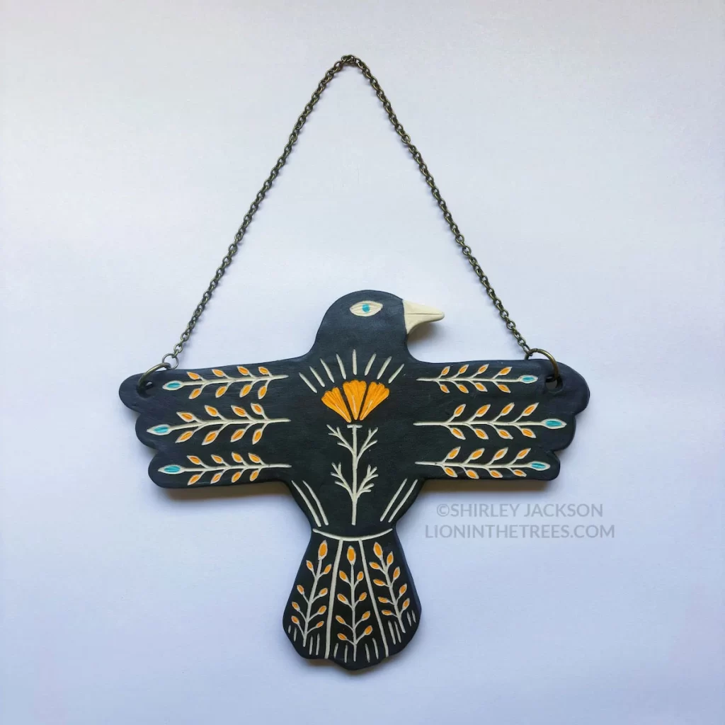 A ceramic wall slab in the shape of a t-posed crow featuring sgraffito floral design elements.