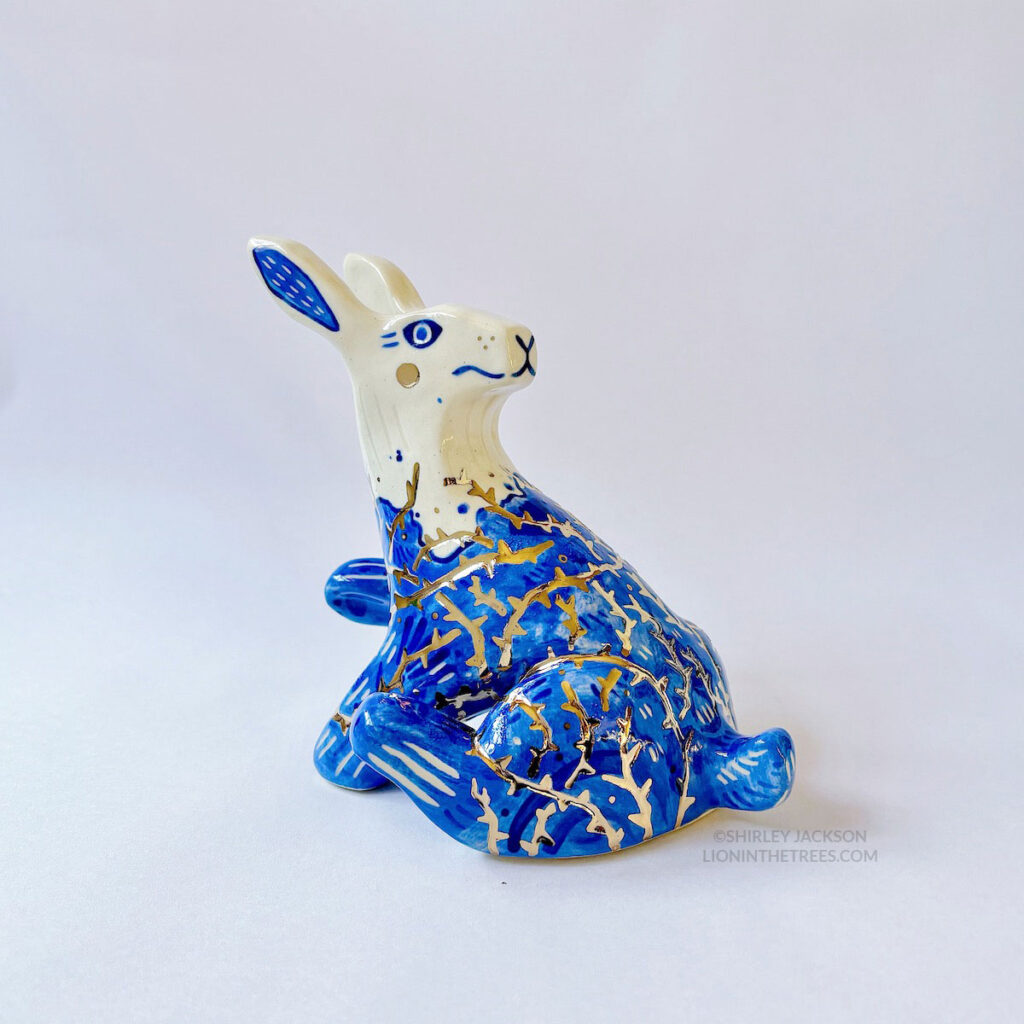 A ceramic sculpture of a rabbit sitting with it's head turned all the way back while also having a paw and a foot raised to give the appearance that they're stuck with tar and thorns all over their body. It's body is done with blue underglaze to mimic blue and white porcelain, with it's lower half covered in the blue underglaze to mimic tar. And gold luster thorns crisscrossing all over it's body.
