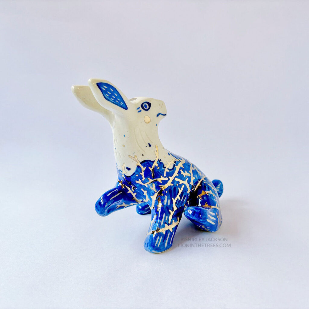 A ceramic sculpture of a rabbit sitting with it's head turned all the way back while also having a paw and a foot raised to give the appearance that they're stuck with tar and thorns all over their body. It's body is done with blue underglaze to mimic blue and white porcelain, with it's lower half covered in the blue underglaze to mimic tar. And gold luster thorns crisscrossing all over it's body.