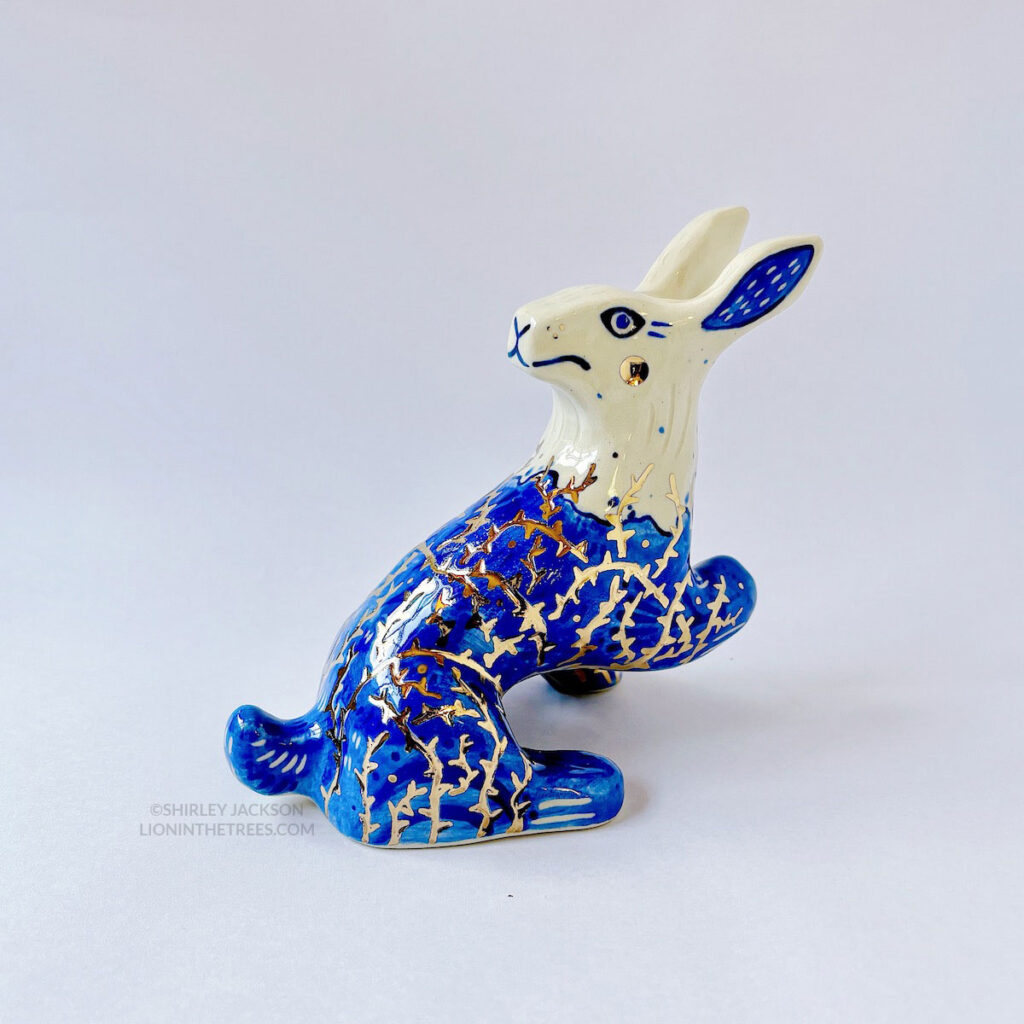 A ceramic sculpture of a rabbit sitting with it's head turned all the way back while also having a paw and a foot raised to give the appearance that they're stuck with tar and thorns all over their body. It's body is done with blue underglaze to mimic blue and white porcelain, with it's lower half covered in the blue underglaze to mimic tar. And gold luster thorns crisscrossing all over it's body.
