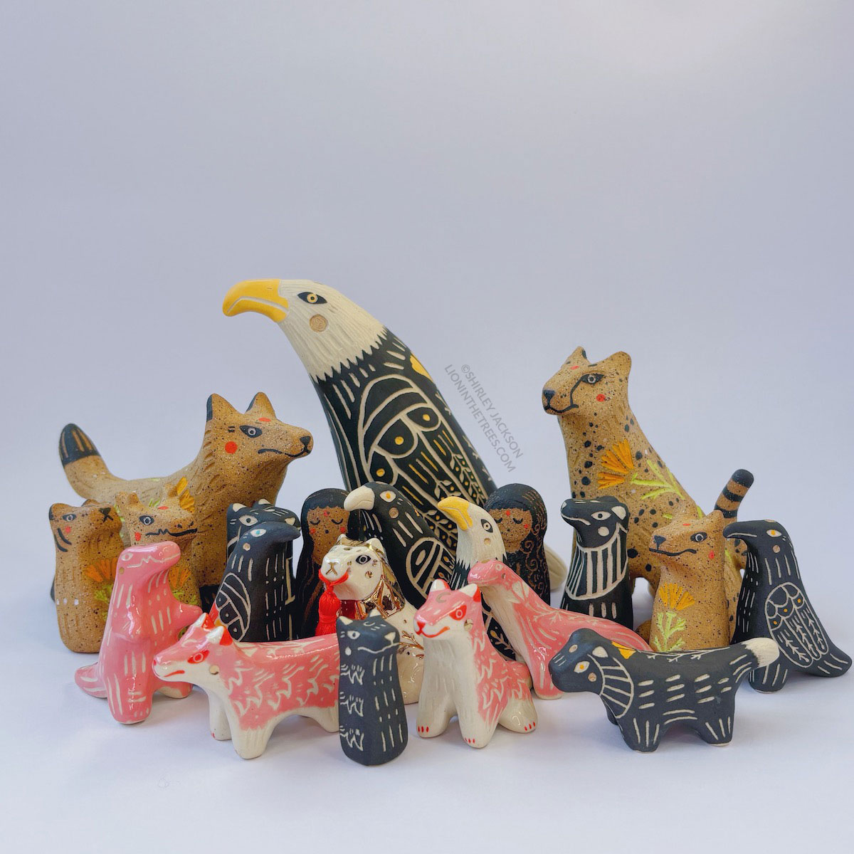 Group photo showing a gathering of ceramic animals. There is a very large eagle in the back along with a brown and speckled fox and cheetah. There are some pink animals such as dinosaurs and wolves, as well as a few birds and human shaped totems as well.