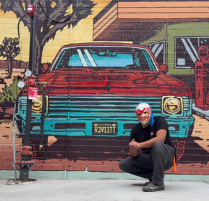 A person wearing a luchador mask posed in front of a beautiful mural of a muscle car.
