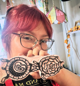 A photo of the artist behind Pen Moon holding up a carved block stamp they made. The design appears to be somewhat floral yet abstract.
