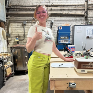 The artist behind Snap Dragon Glass smiling and giving a thumbs up in a workshop space.